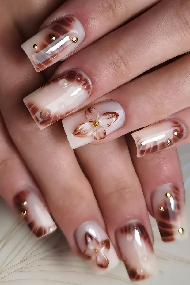 Elegant floral nail art design with brown and cream tones, featuring gold studs and intricate patterns for a trendy look.