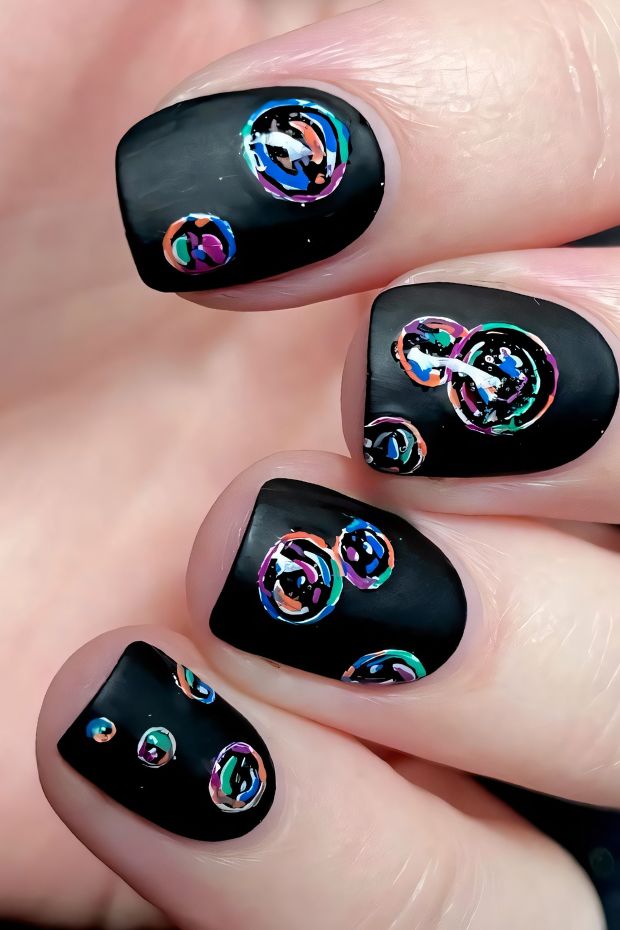 Black nails adorned with colorful bubbles in vibrant hues and a matte finish, showcasing unique nail art design.