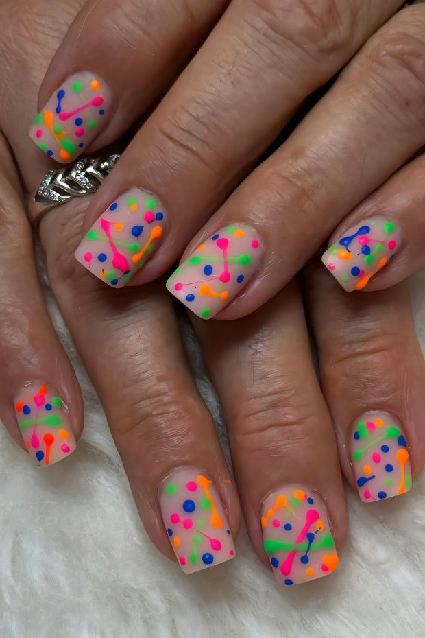 Short nails with a nude base and vibrant neon splatter in pink, green, orange, and blue.