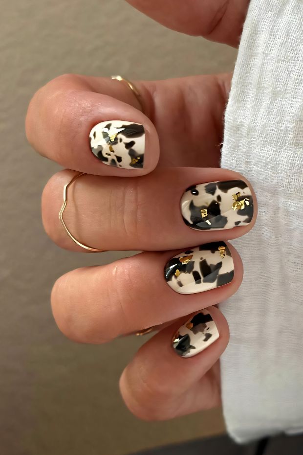 Stylish short nails with cow print design in black and white, complemented by gold foil accents for a trendy look.