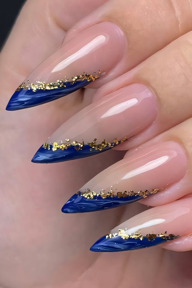 Stiletto nails with navy blue tips and gold flakes on a nude base for a modern nail art design.