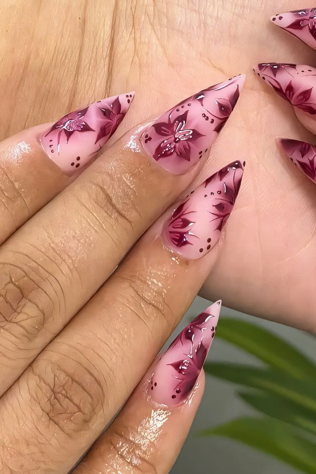Stiletto nail design featuring burgundy floral patterns with glitter accents on a nude base.