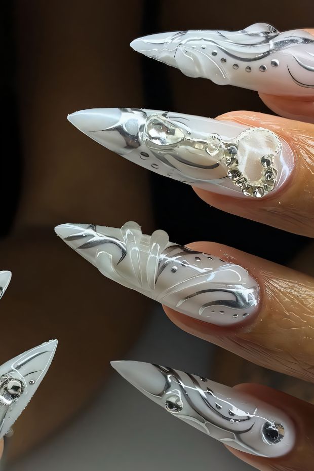 Stiletto nails with white base and intricate silver designs adorned with gems and embellishments.