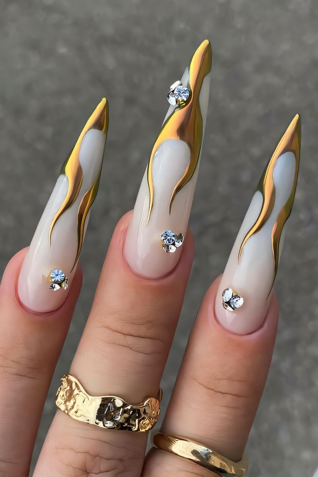 Stiletto nails with gold flames and rhinestones on a nude base, showcasing a glamorous nail art design.