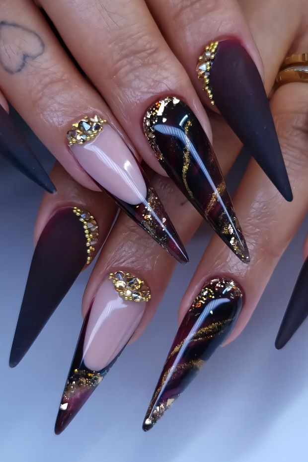 Glamorous stiletto nails in burgundy, black, and gold with intricate detailing and stunning embellishments.