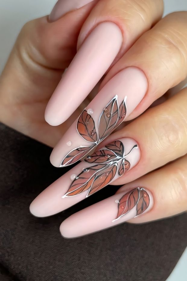 Matte nude nails with delicate leaf designs and metallic accents, offering a chic and modern nail art style.