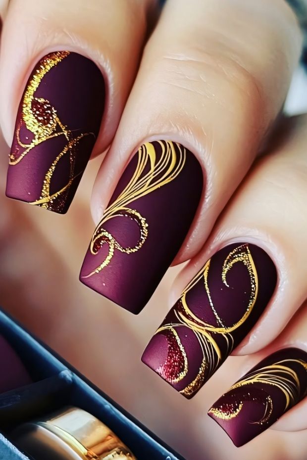Matte burgundy nails with intricate gold designs and elegant swirls, showcasing a trendy nail art style.