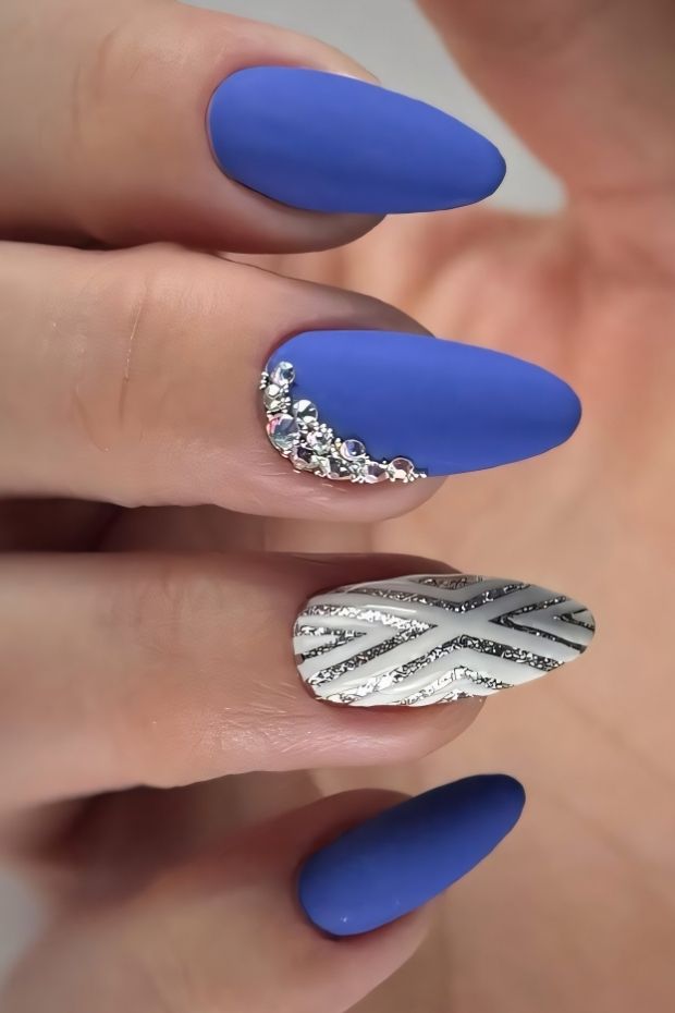 Matte blue nails with silver rhinestone accents and a detailed design on the ring finger.