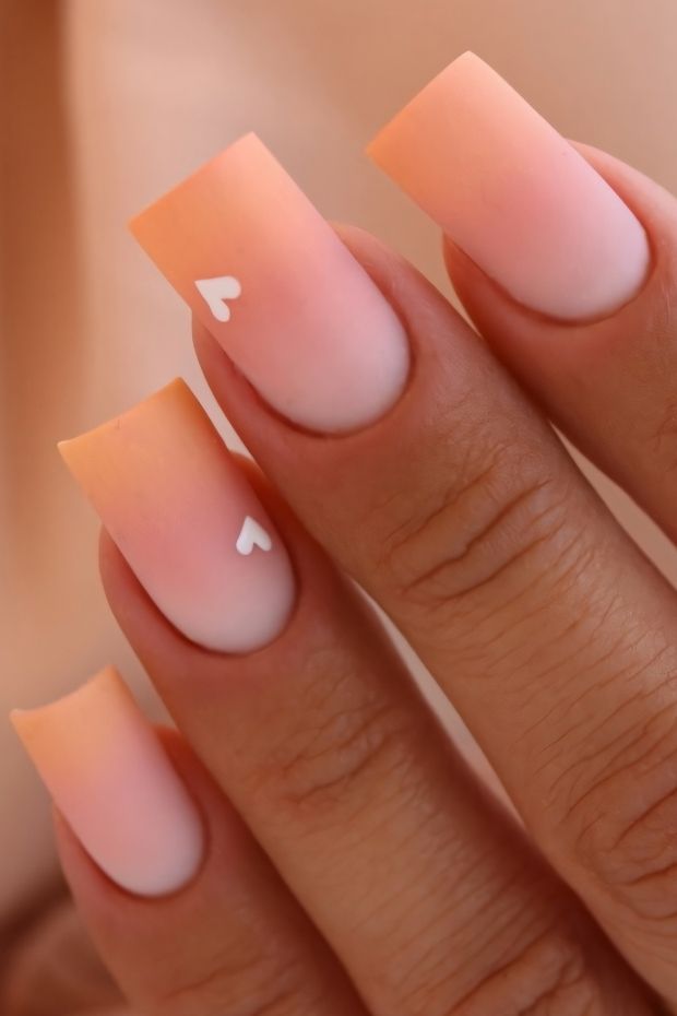 Stylish matte ombre nails in peach to nude with white heart accents.